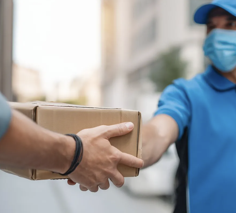 Delivery person in blue uniform and mask handing cardboard package to customer