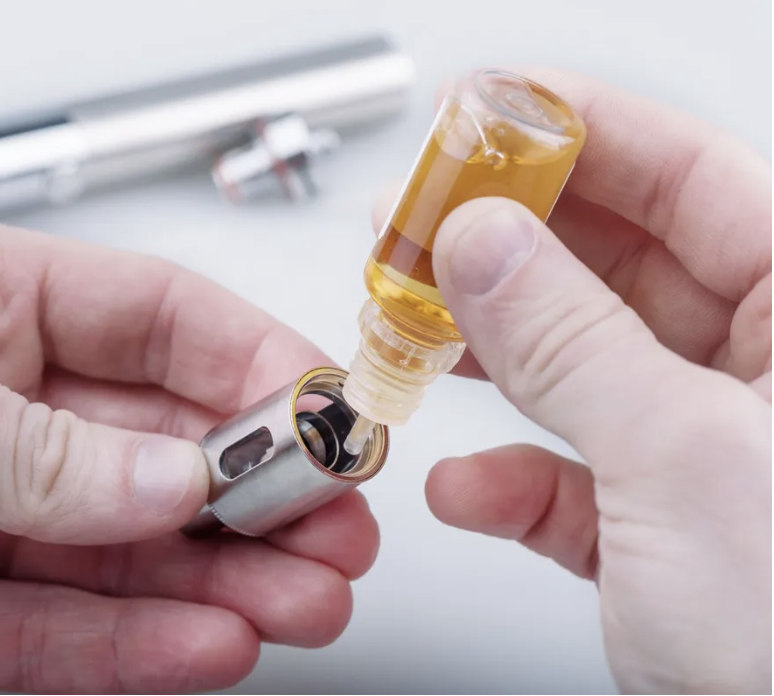 Person holding a vape cartridge with golden liquid being inserted into a silver battery device